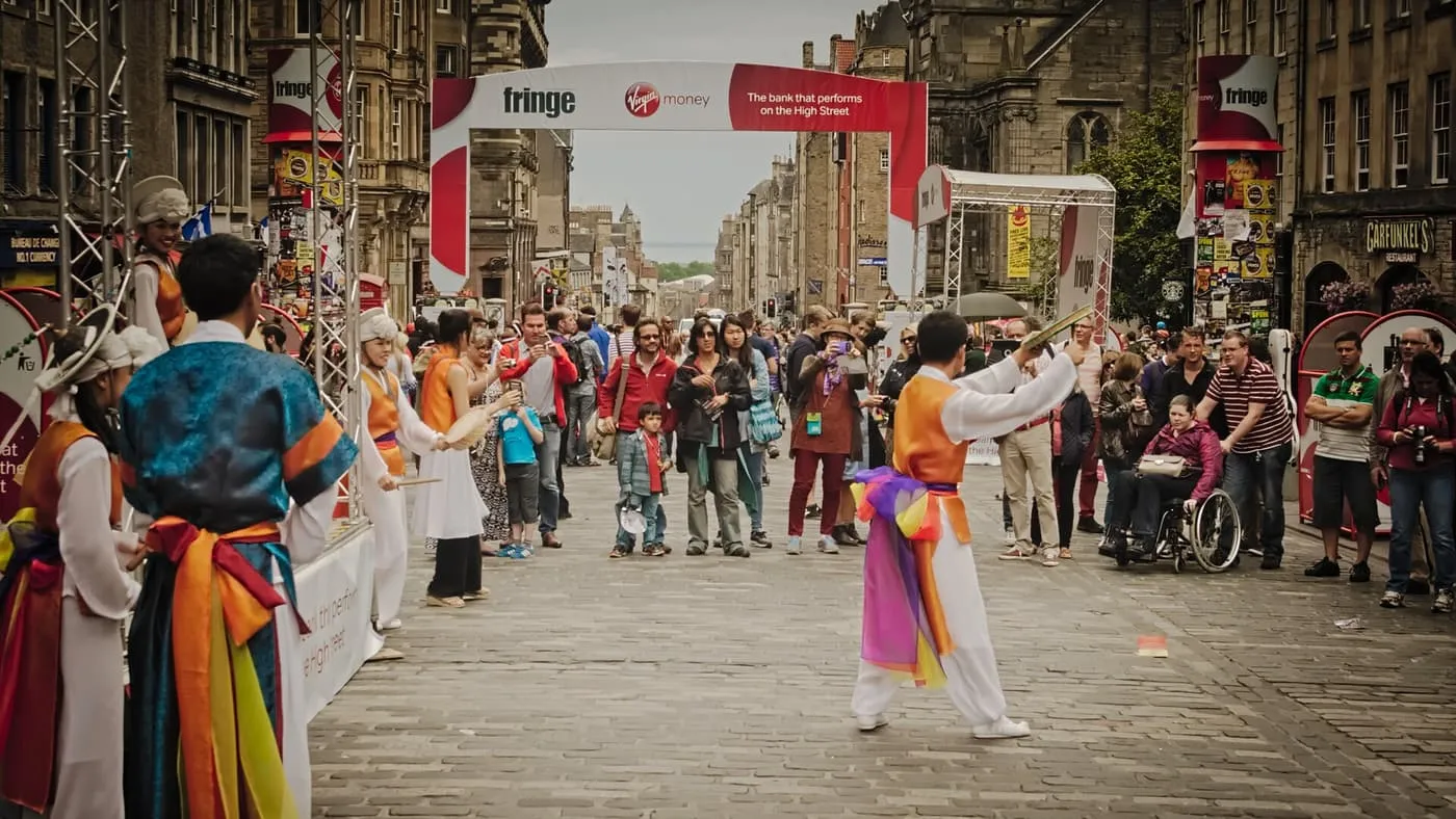 A performer in orange and white dancing on the cobbles as an audience gathers, under a Fringe banner arching over the High Street