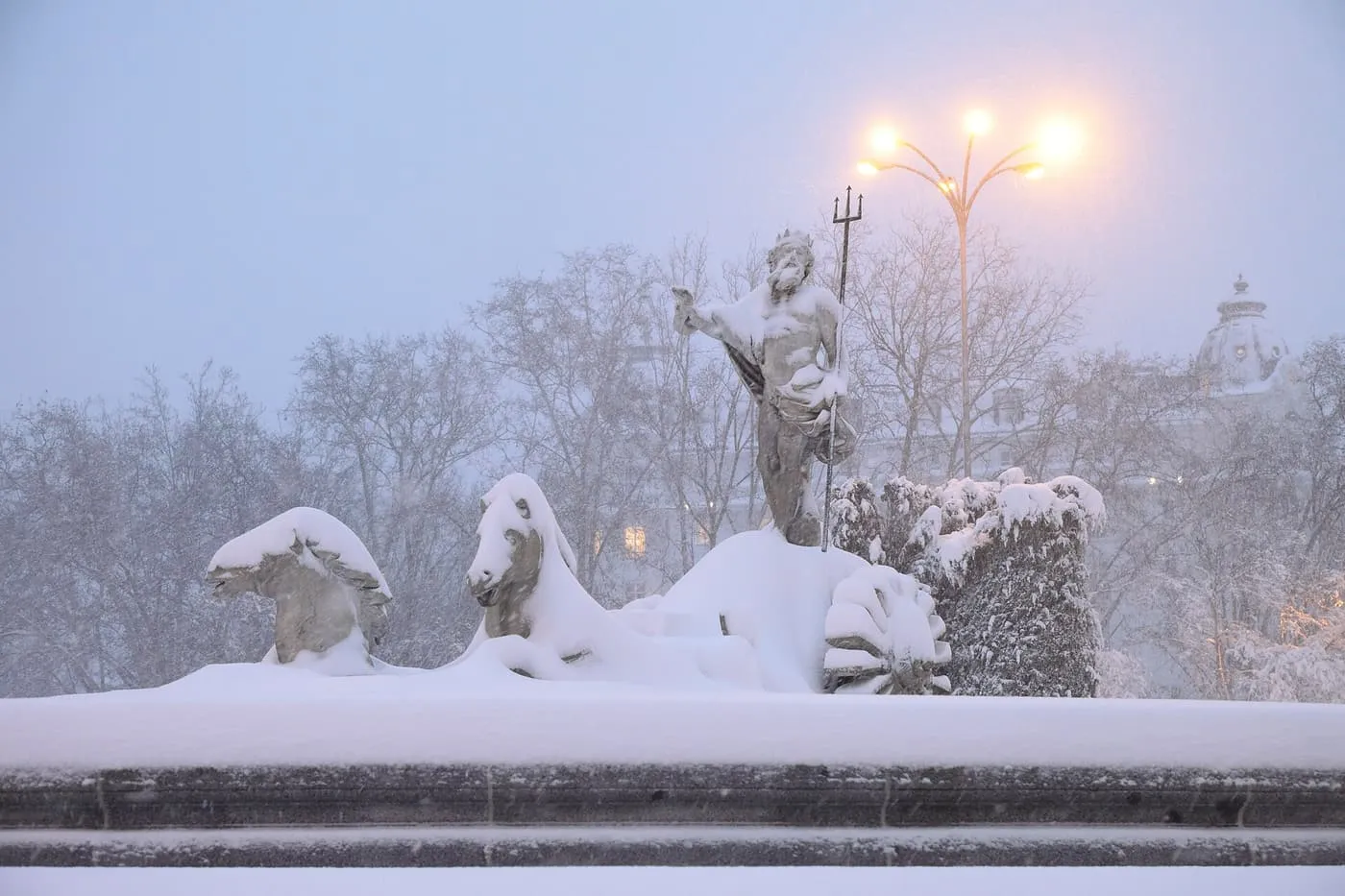 The statue of Neptune and his sea horses on the Fuente de Neptuno, covered in snow, with a street lamp glowing behind