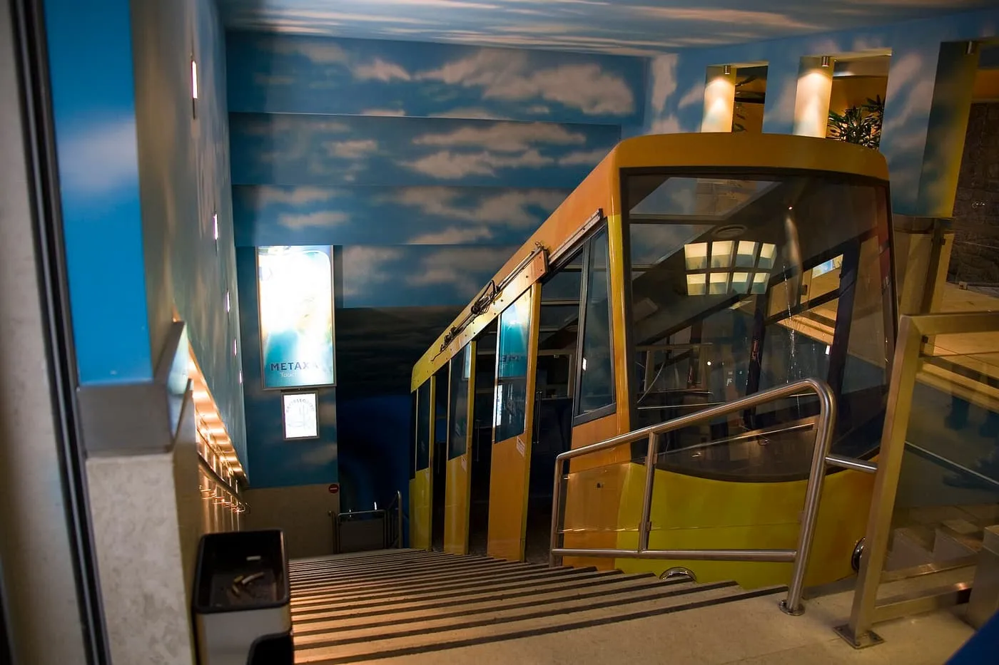 A yellow funicular car in a station with a sky-painted ceiling, beside a flight of steps
