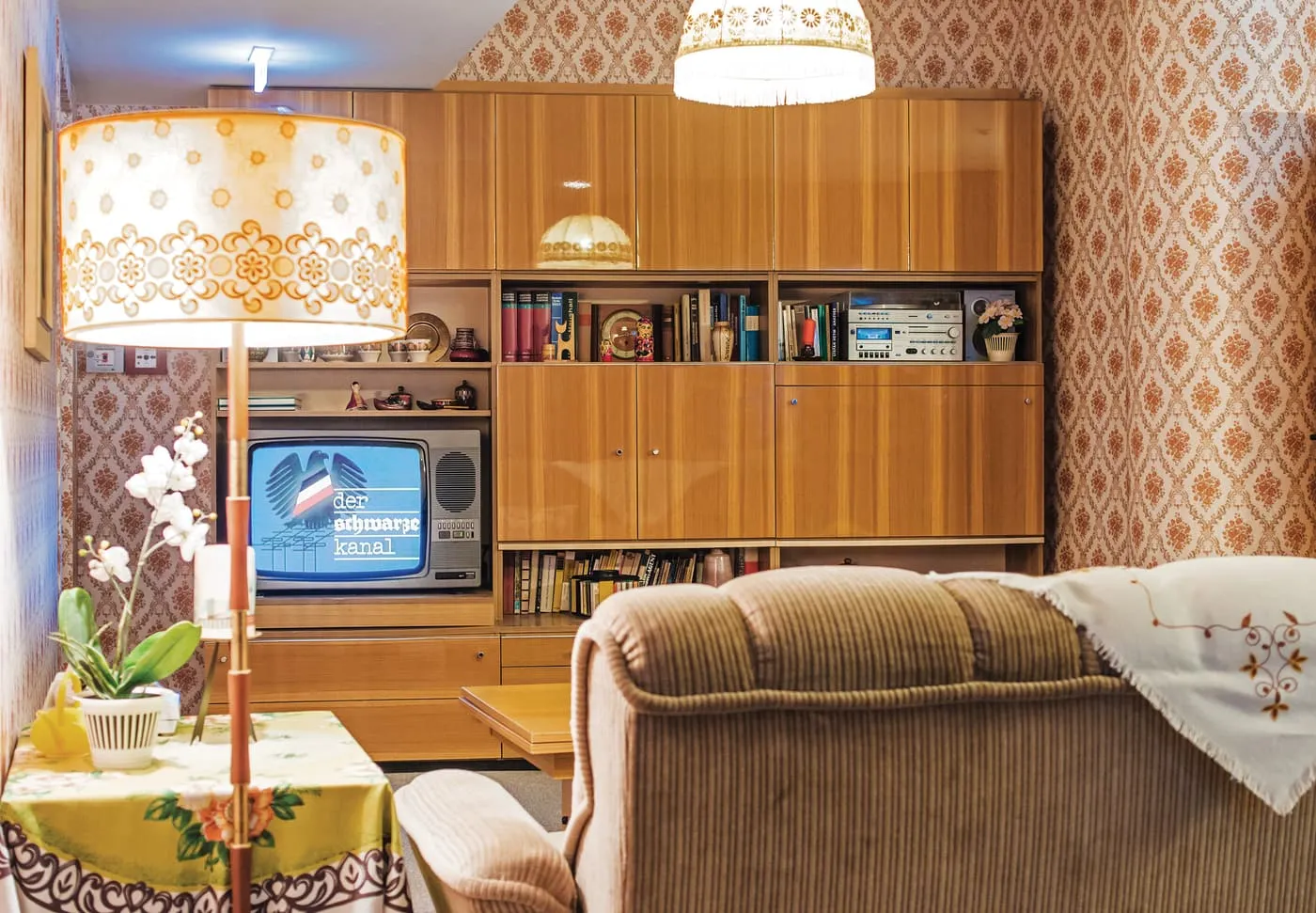 A furnished East German living room in the museum, with patterned wallpaper, a wooden wall unit, a television and a corduroy armchair