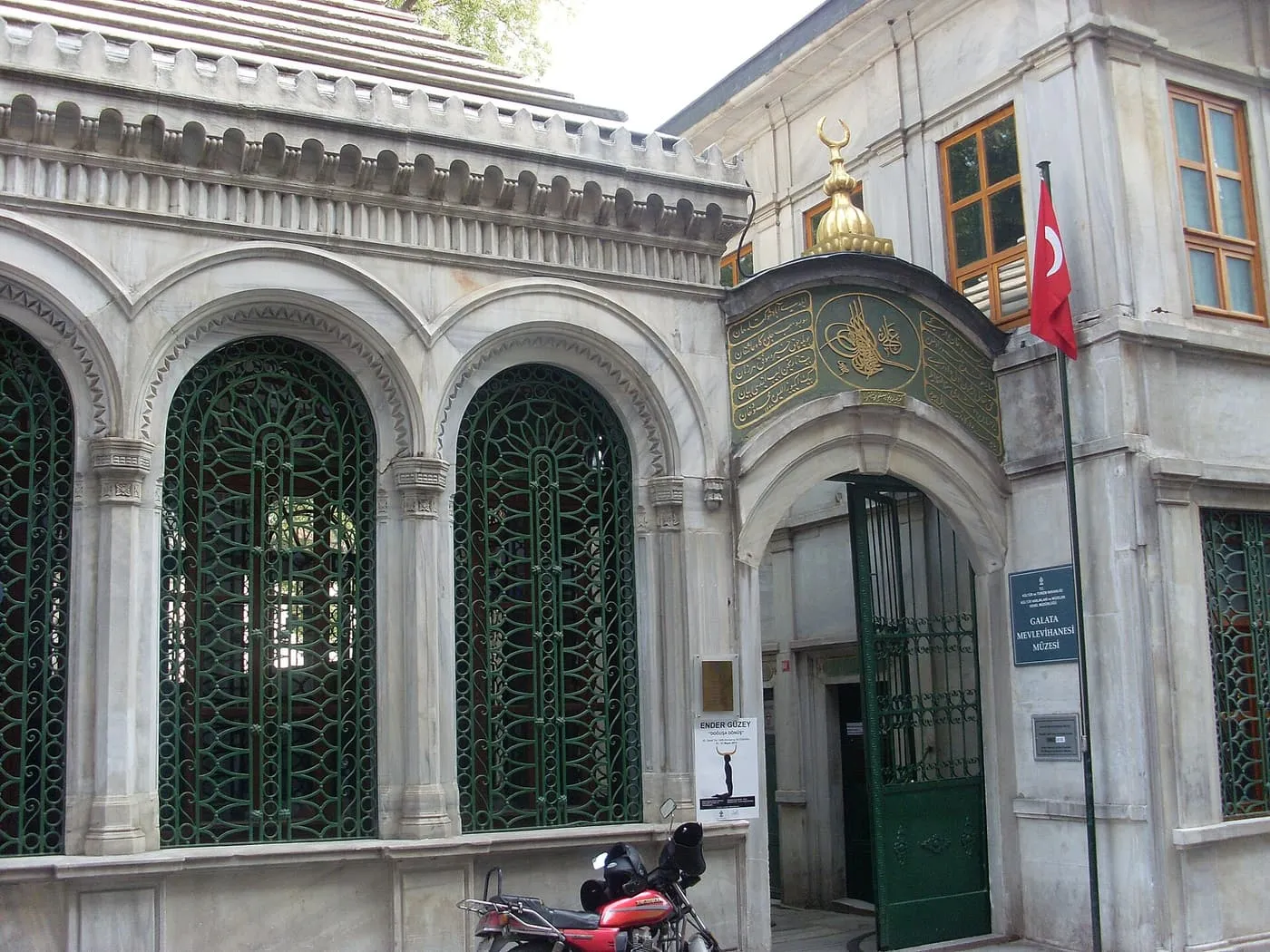 The marble entrance of the Galata Mevlevi Lodge in Beyoğlu, with a green and gold calligraphic panel over the arched gate and a Turkish flag beside it