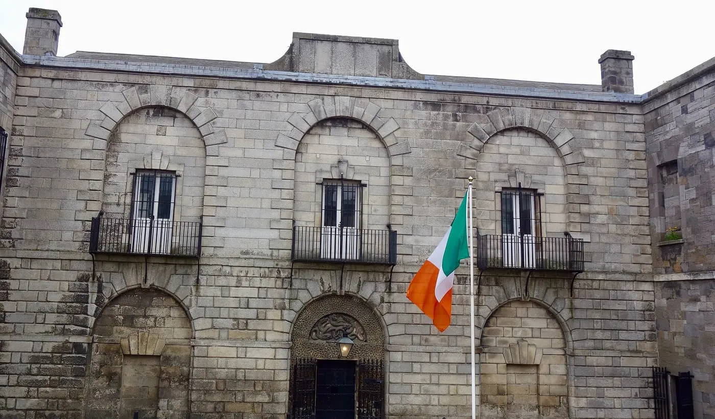 The stone front of Kilmainham Gaol with three arched windows with balconies, an arched doorway and an Irish flag