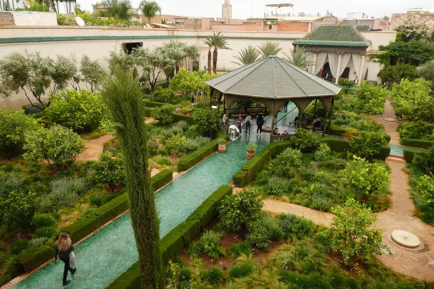 A green-tiled water channel between clipped hedges leads to a pavilion in a walled garden, seen from above, among orange and olive trees