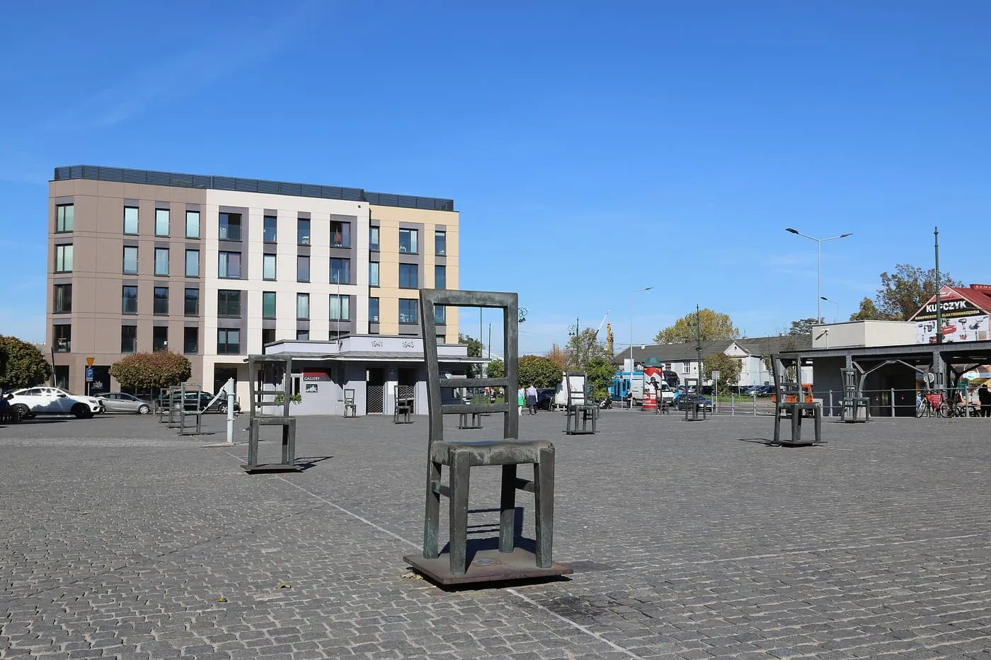 Empty bronze chairs standing on the paving of Ghetto Heroes Square, with apartment blocks behind and a blue sky