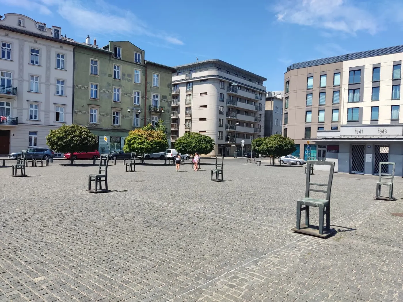 Empty bronze chairs standing on the paving of plac Bohaterów Getta, with apartment blocks behind