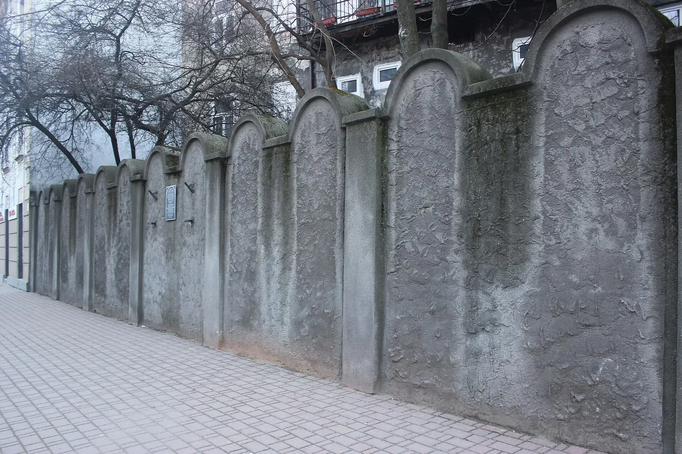 A grey plastered wall with rounded tops like gravestones running along a paved pavement