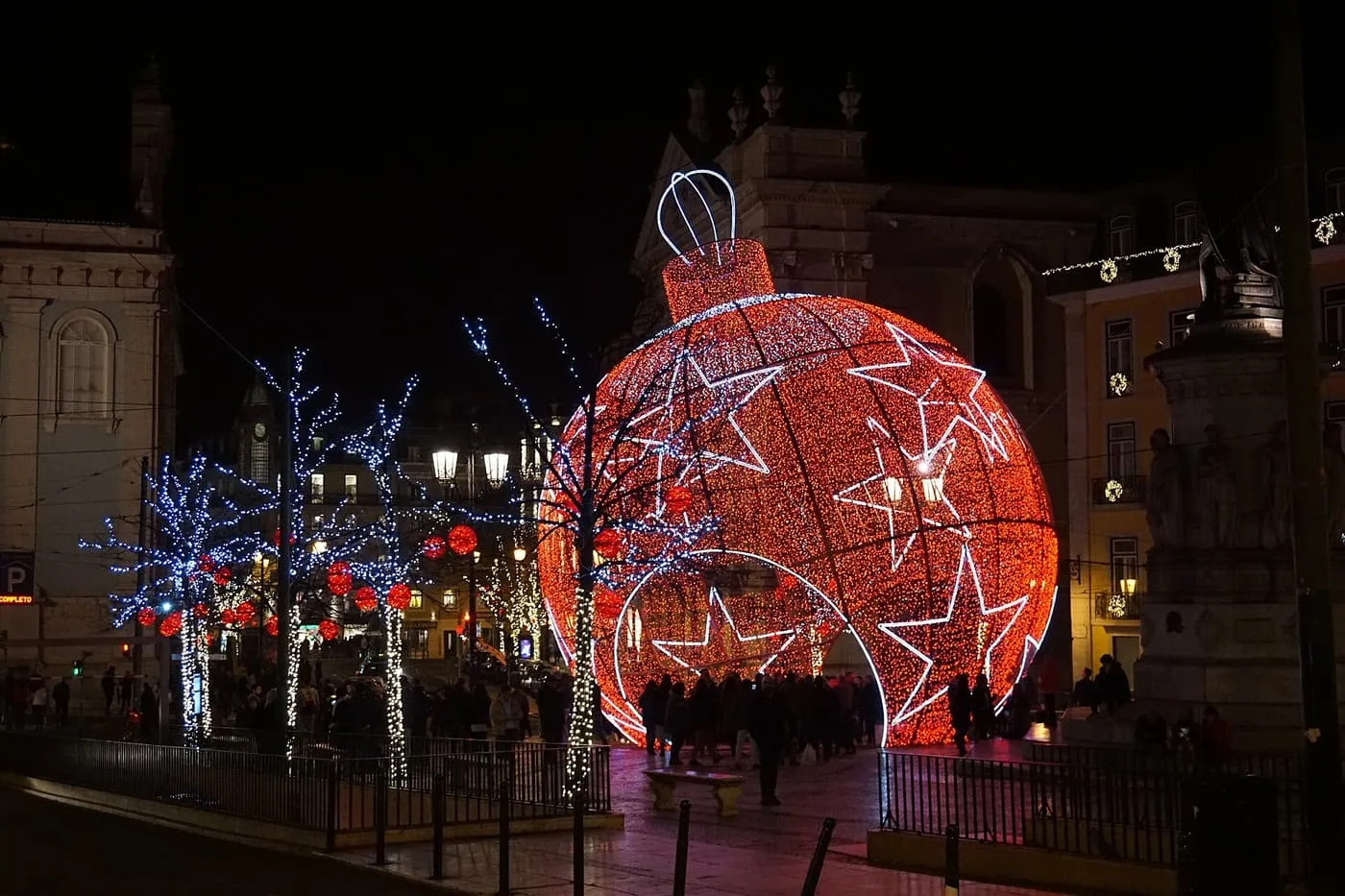 A giant red bauble made of lights standing among trees strung with white and red lights, with a crowd at its base at night