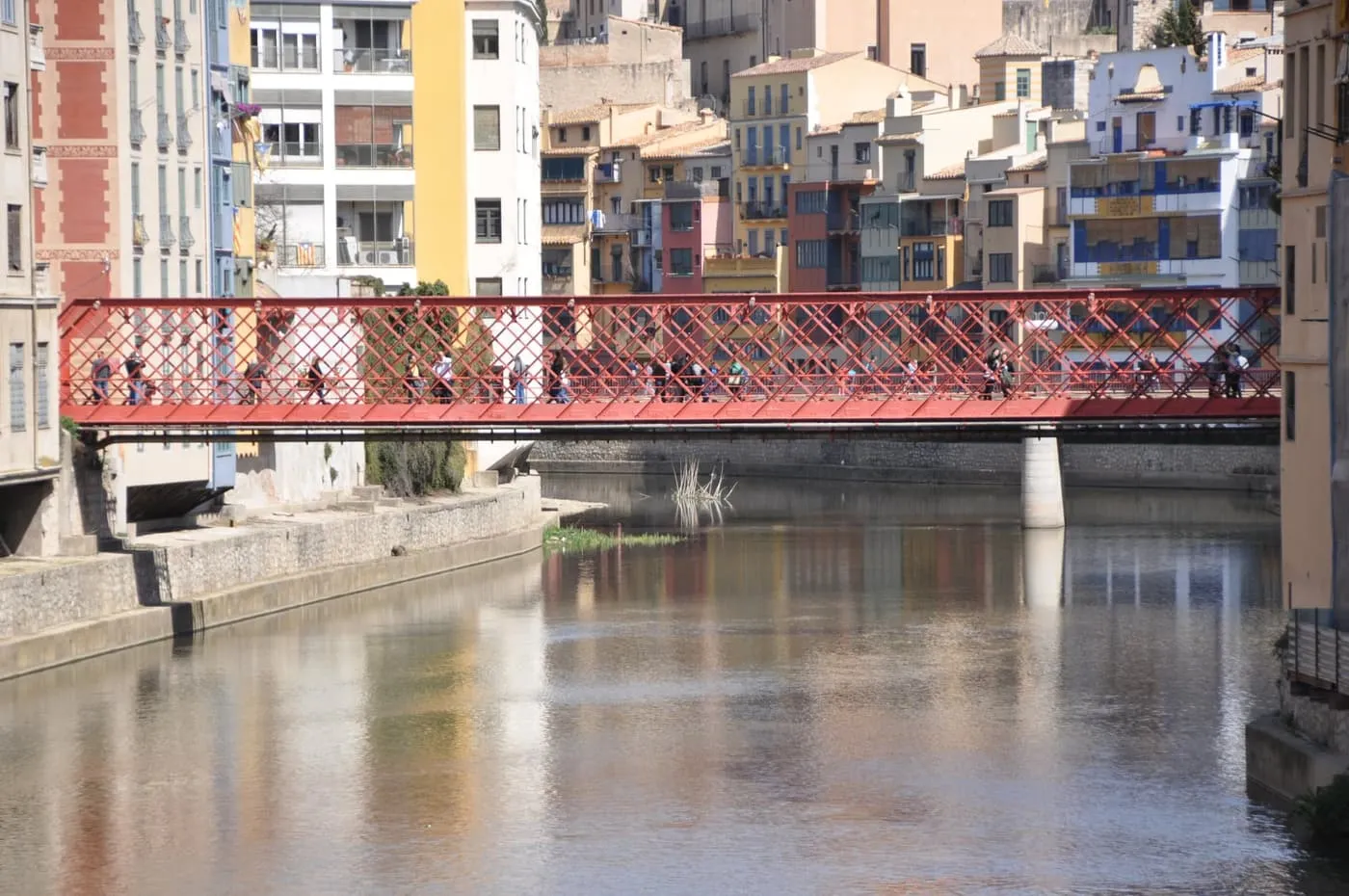 A red iron lattice footbridge over the River Onyar in Girona, with tall painted houses along the far bank