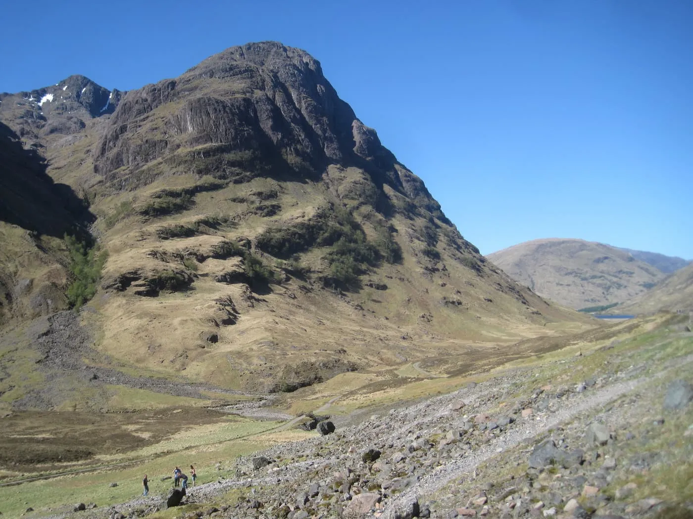 A steep-sided mountain at the entrance to Glen Coe under a blue sky, with walkers on the scree below