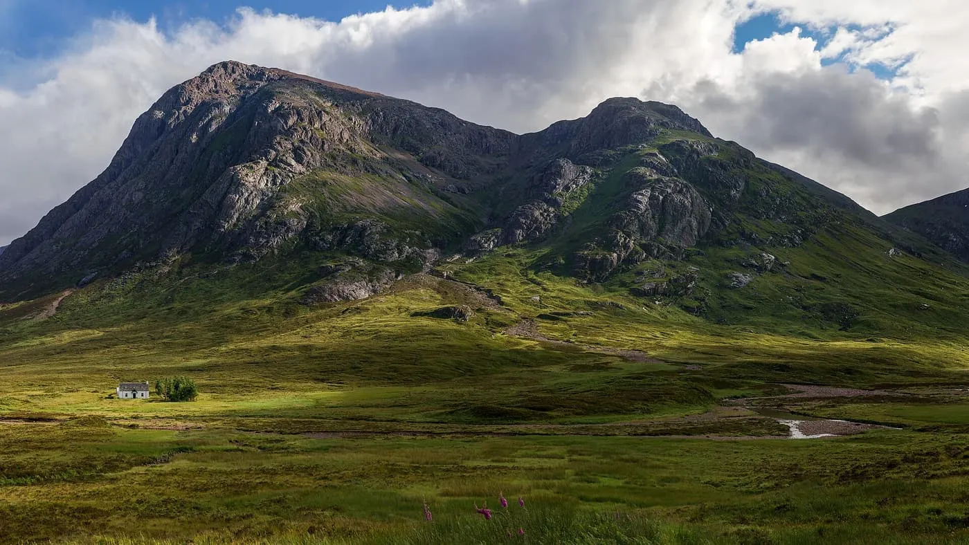 The rocky peaks of Buachaille Etive Mòr rising above Glencoe moorland, with a small white cottage in the sunlit foreground
