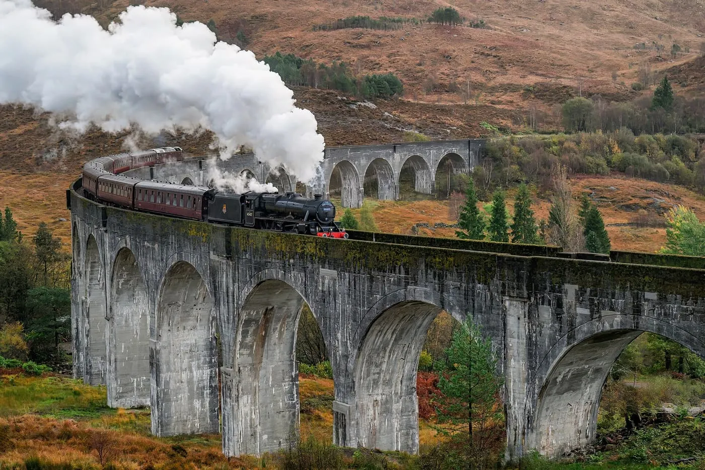 A steam train with a plume of white smoke crossing the curved stone arches of the Glenfinnan Viaduct