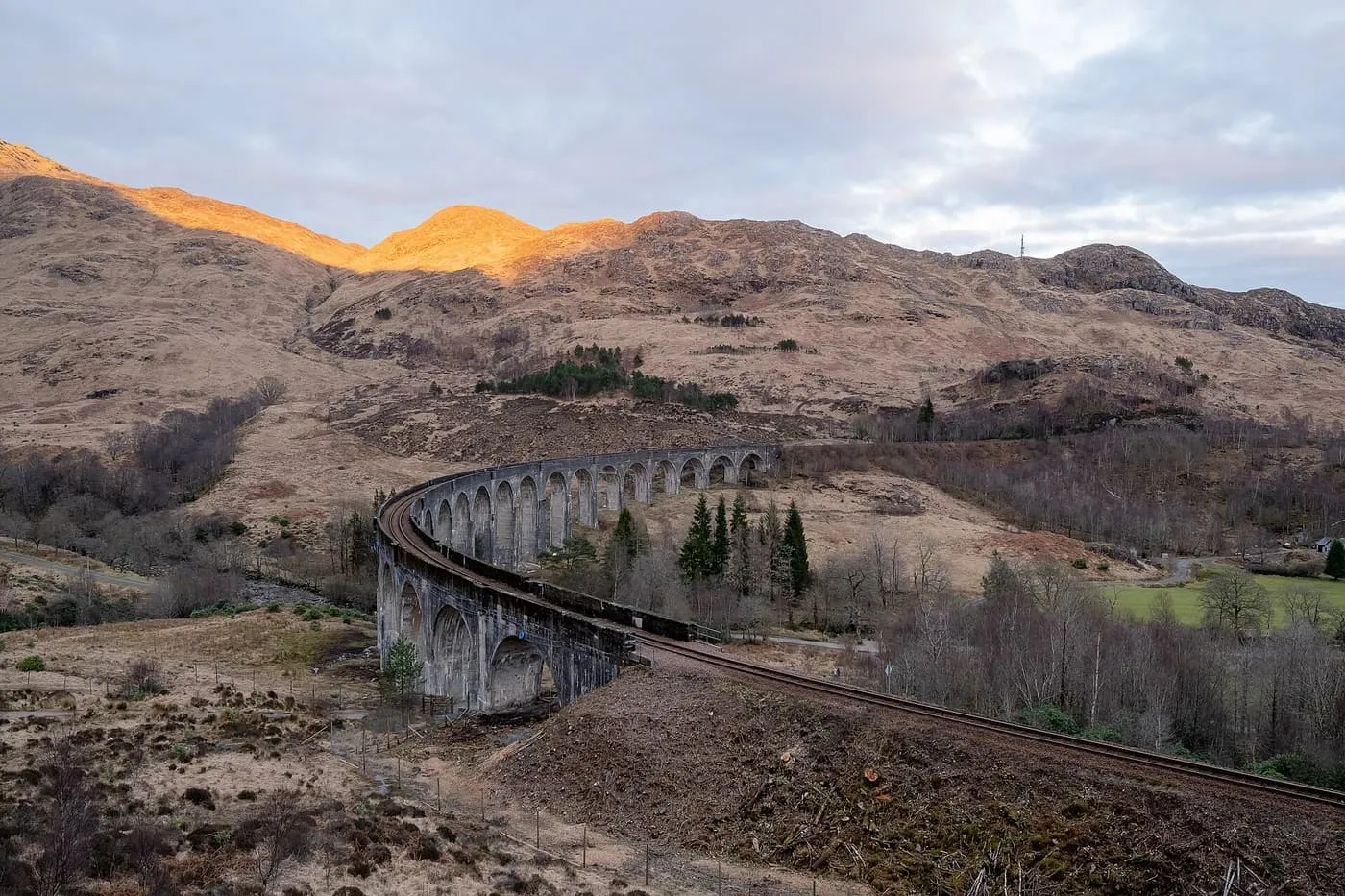 The curved Glenfinnan Viaduct crossing a glen, with sunlit hilltops behind