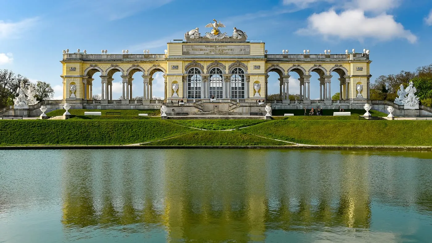 The arcaded Gloriette at Schönbrunn on its hill above a pond, under a blue sky with a lawn in the foreground