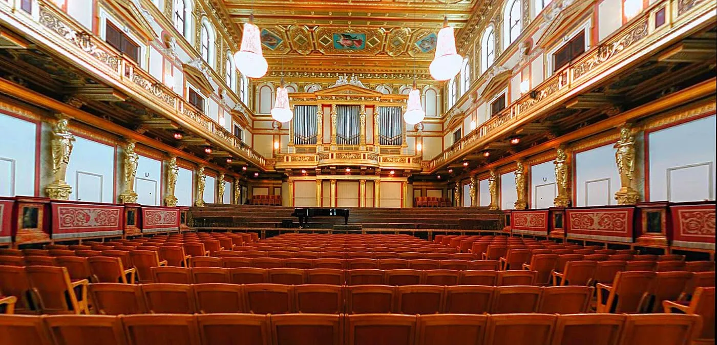 The Golden Hall of the Musikverein, a long hall with a gilded coffered ceiling, gilded figures along the walls, an organ at the far end and rows of red seats