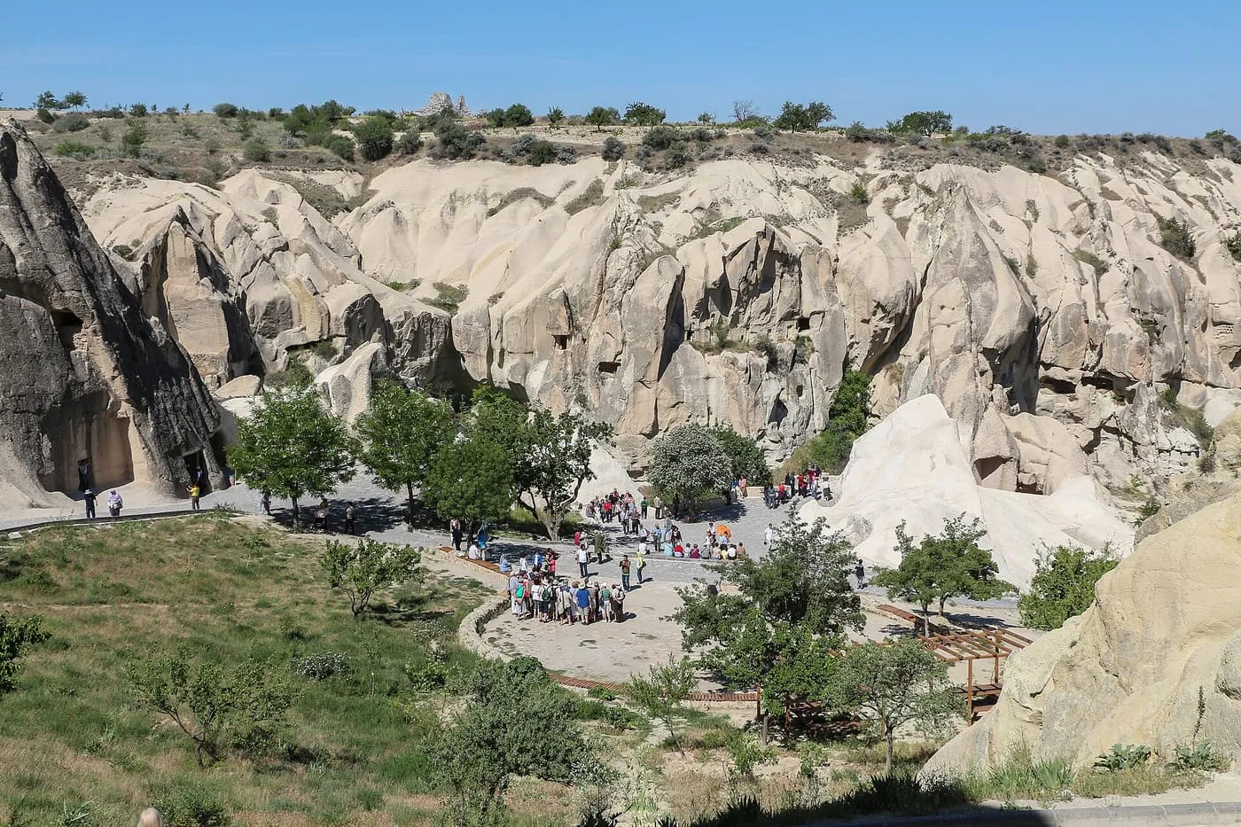 A valley of pale rock cliffs at the Göreme Open Air Museum, with a visitor path and small groups of people among the trees