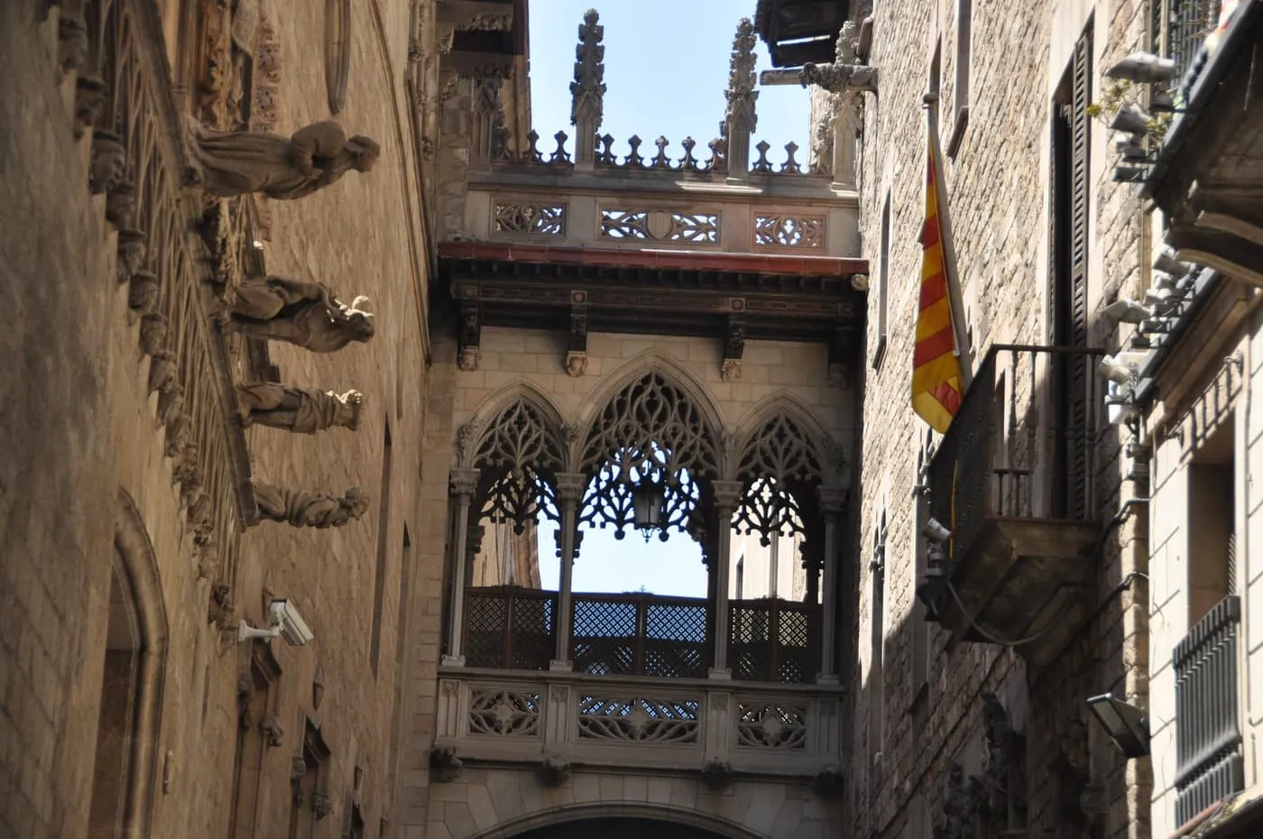 A carved neo-Gothic stone bridge spanning a narrow lane between old stone buildings in the Gothic Quarter, a Catalan flag hanging from a balcony
