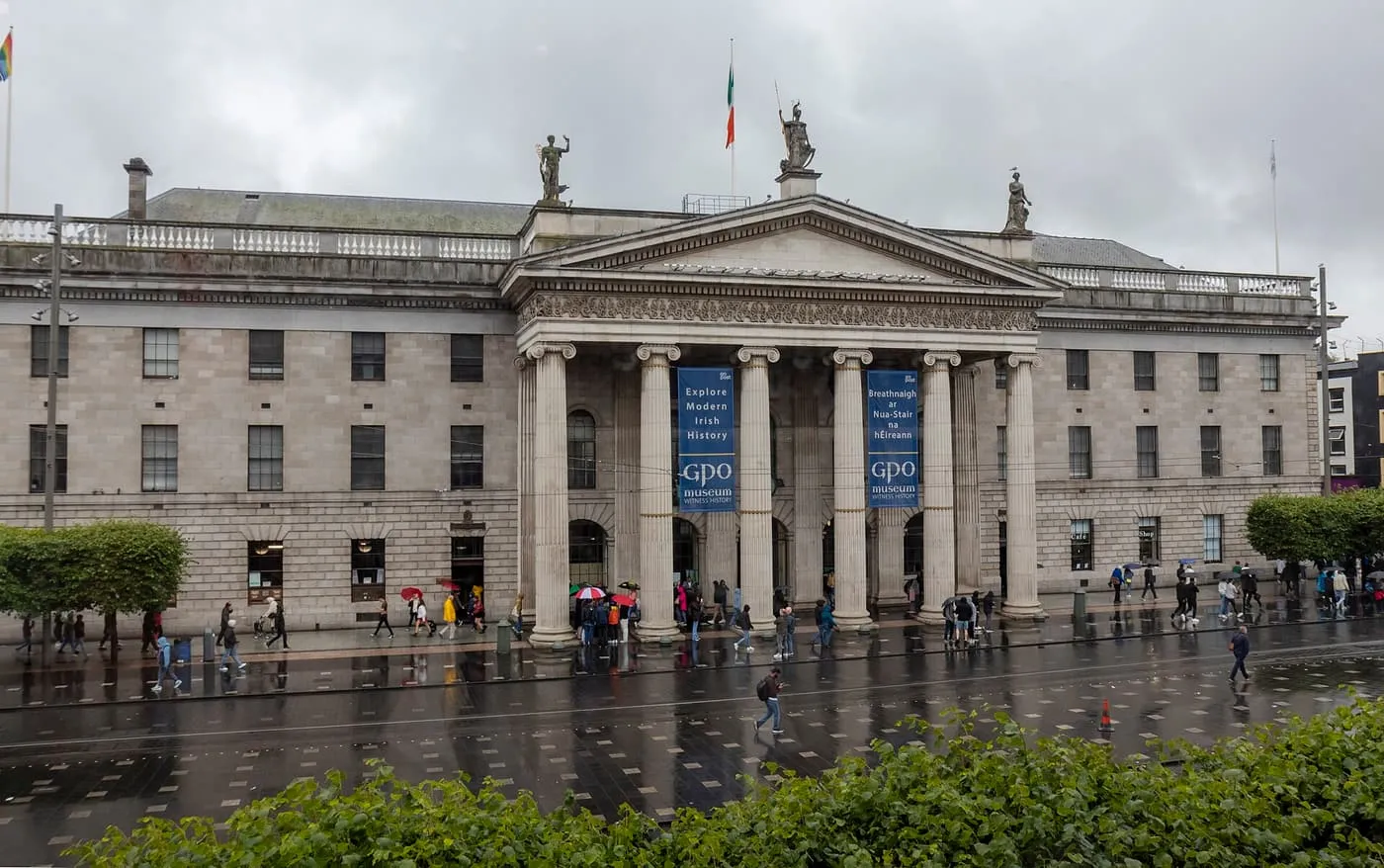 The columned Ionic portico and grey stone front of the General Post Office on O'Connell Street, with an Irish flag on the roof and people crossing the wet pavement