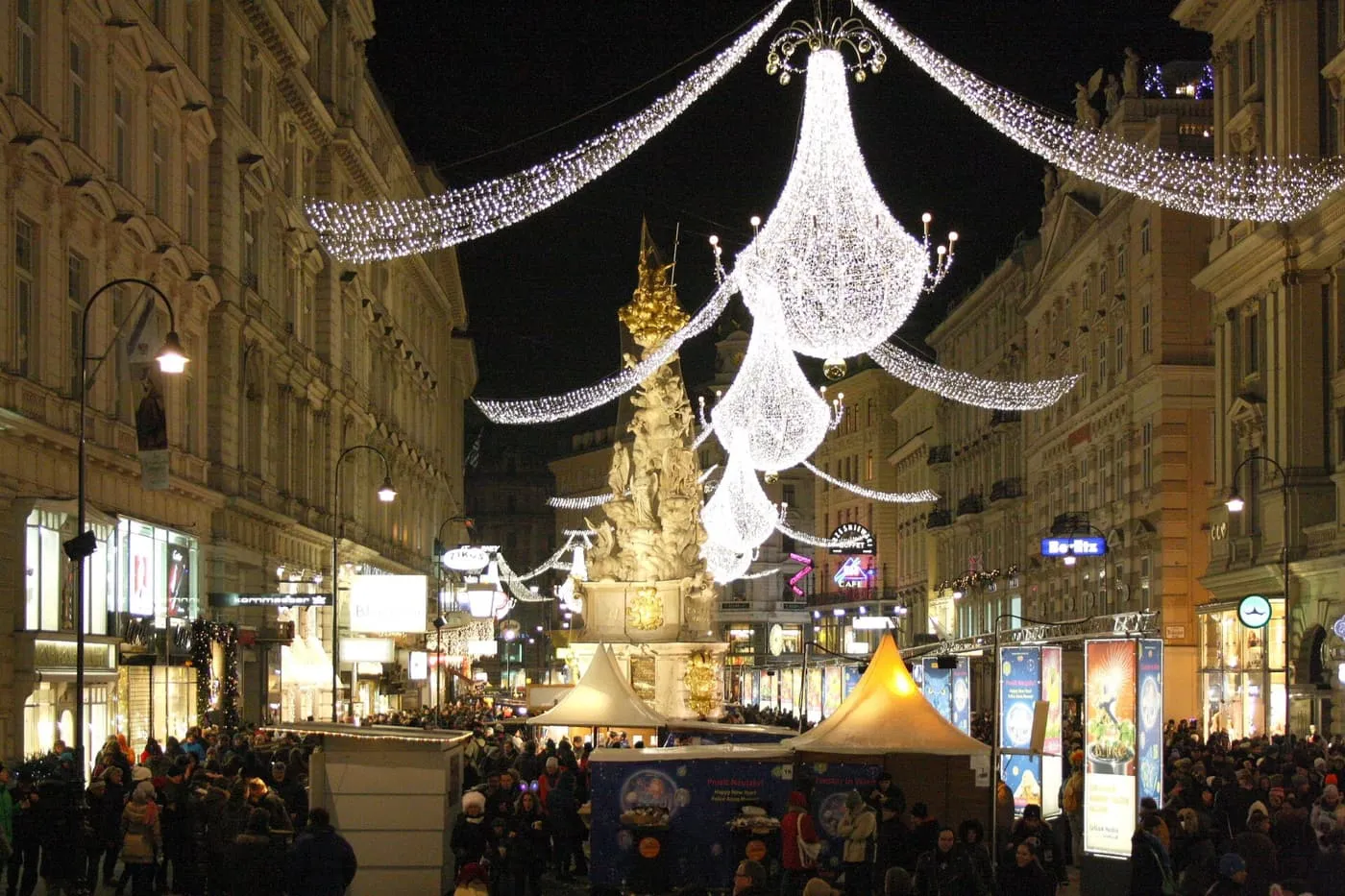 The Graben at night, with the Plague Column lit gold under hanging chandeliers of lights and a crowd among the market stands
