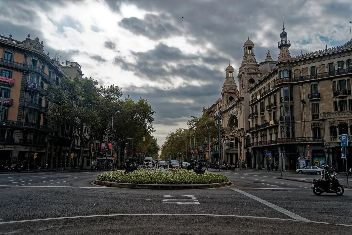 Gran Via de les Corts Catalanes in the Eixample: a wide tree-lined avenue with a roundabout garden and ornate stone buildings under a cloudy sky