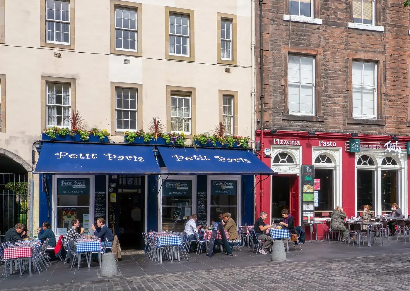 Diners at checked-cloth tables under the blue awnings of Petit Paris and beside a red-fronted pizzeria on the Grassmarket
