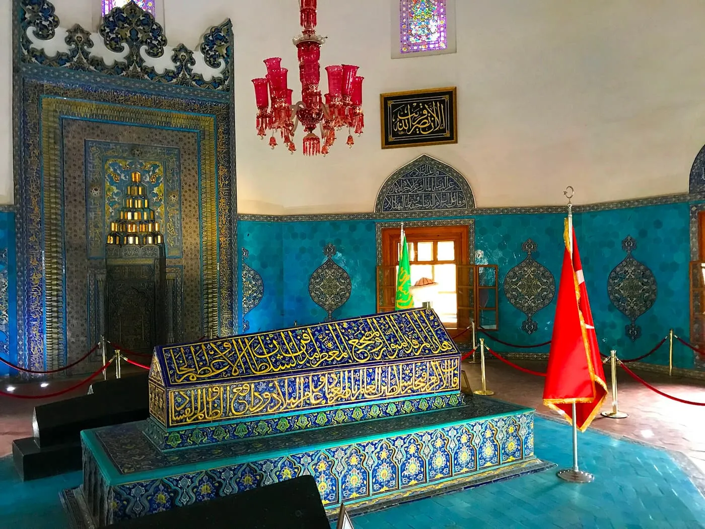 Inside the Green Tomb in Bursa: turquoise hexagonal tiles line the walls around a tile-covered sarcophagus, with a red glass chandelier overhead