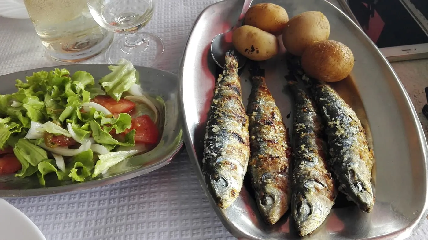 Four grilled sardines on a metal dish with boiled potatoes, beside a bowl of salad