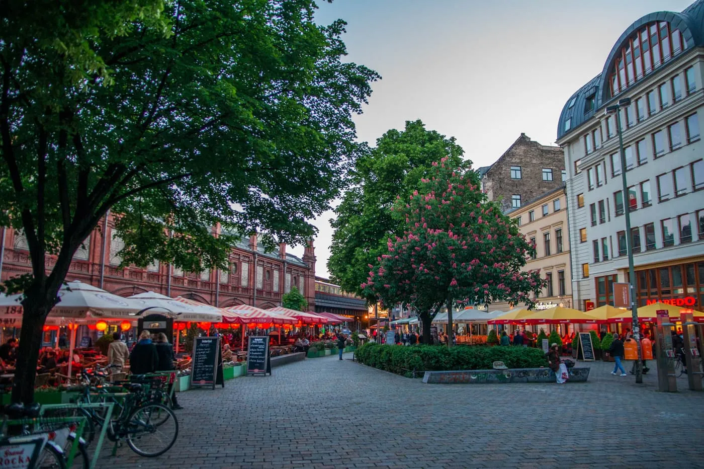 Restaurant terraces under parasols along a cobbled square beside the red-brick S-Bahn viaduct at Hackescher Markt, with a flowering tree, at dusk