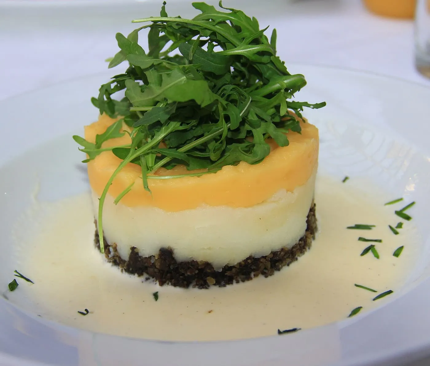 Haggis, mashed potato and mashed neeps stacked in layers on a white plate, with cream sauce around them and a handful of rocket on top