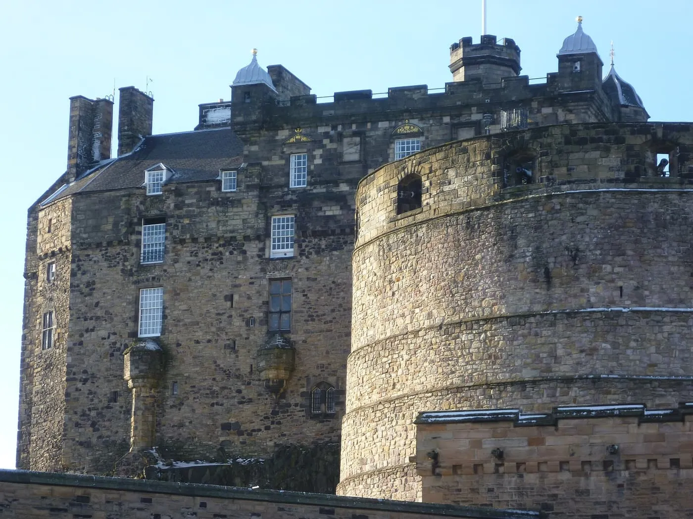The round stone tower of the Half Moon Battery in front of the tall Palace Block of Edinburgh Castle, with a blue sky behind