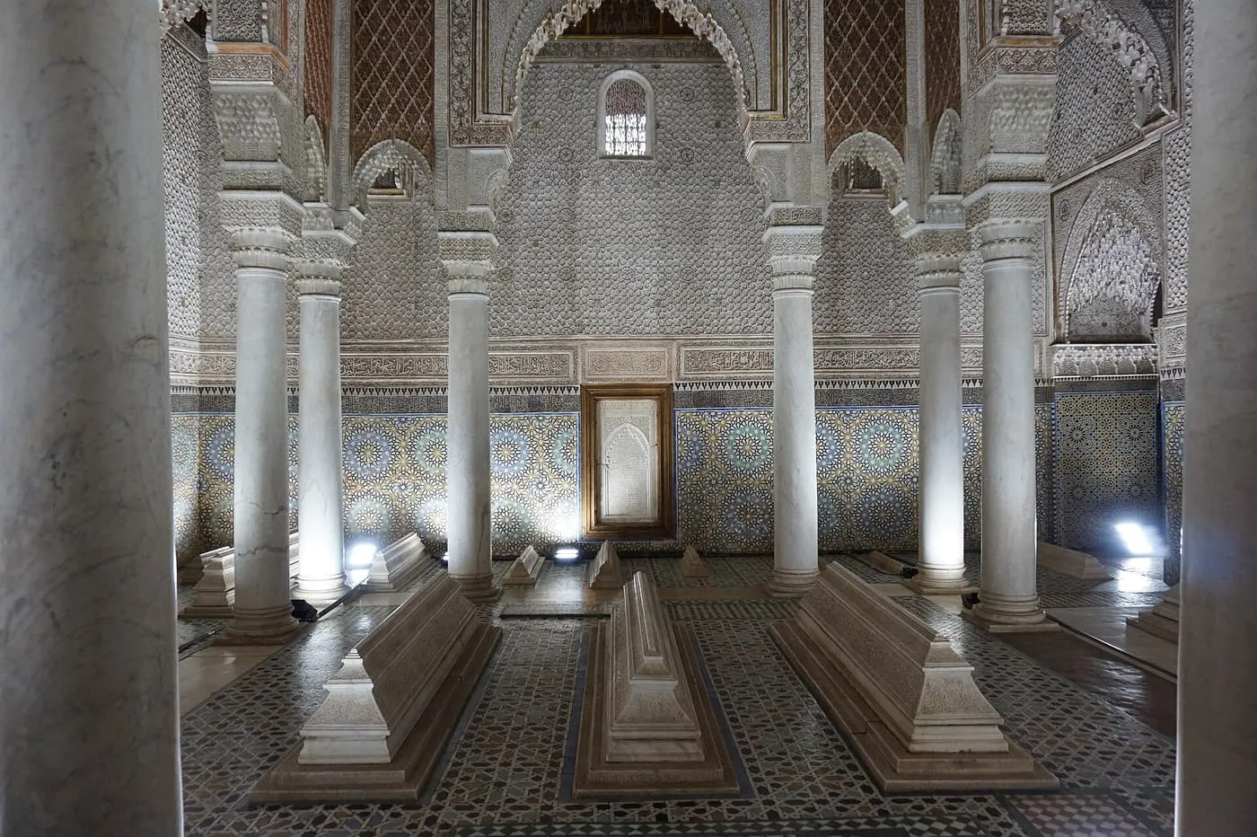 Marble columns and white tomb slabs in the Hall of Twelve Columns at the Saadian Tombs, with carved plaster and tilework on the walls behind