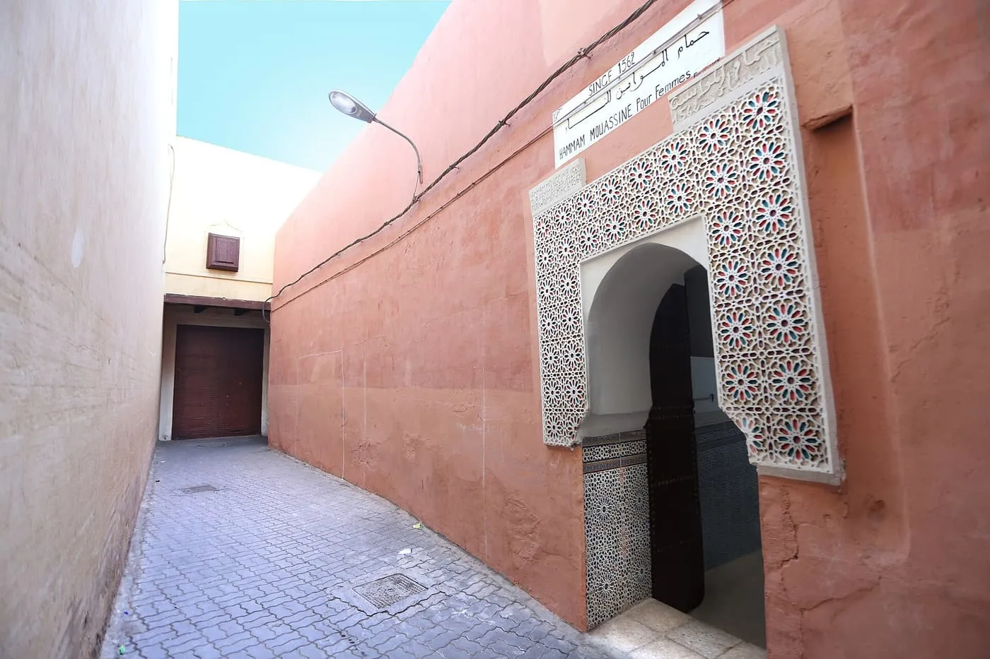 A narrow paved lane with a carved plaster doorway in a pink wall, under a sign for Hammam Mouassine