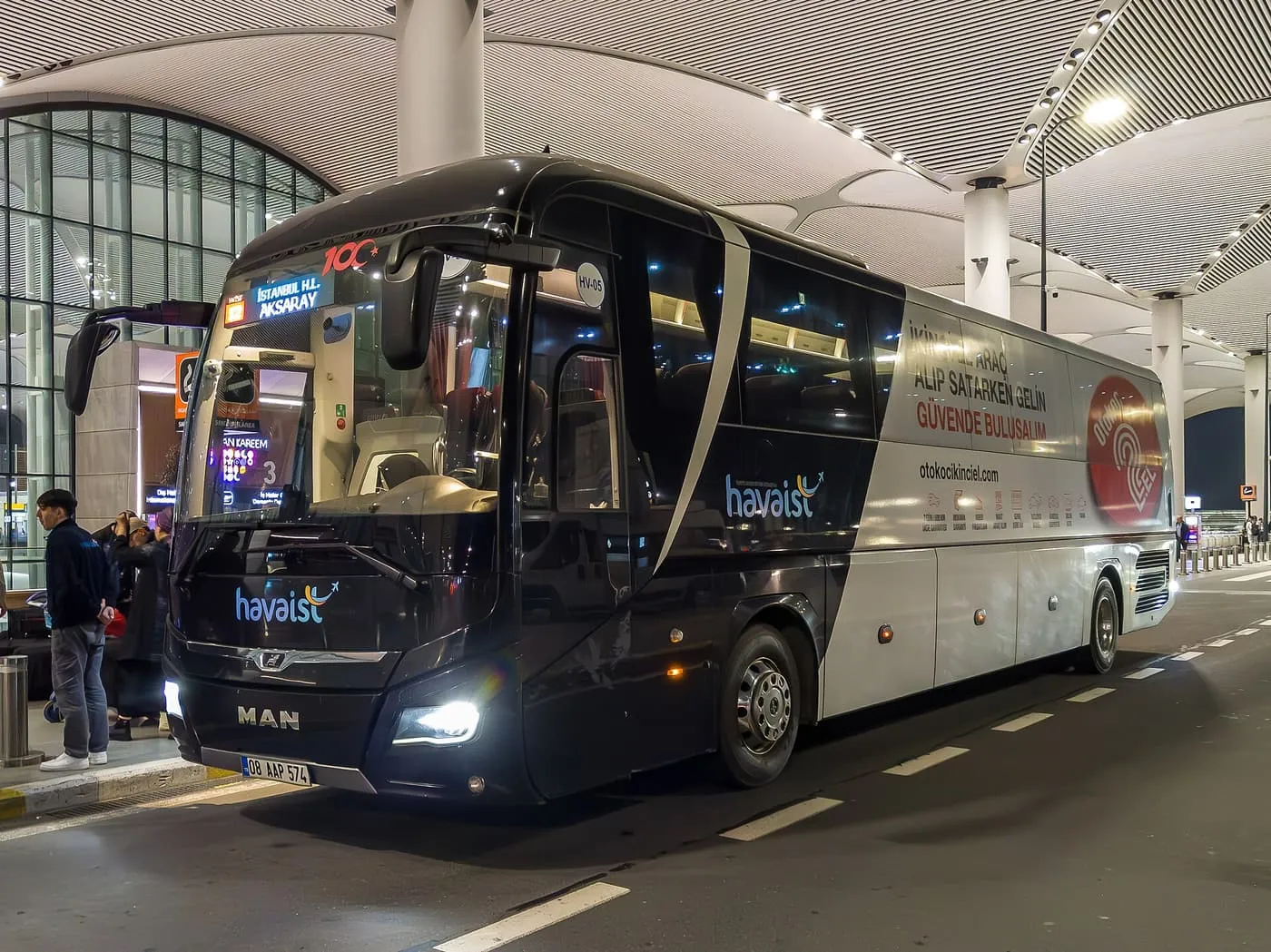 A black and silver Havaist coach with an Aksaray destination board, parked at the kerb under the terminal roof at night
