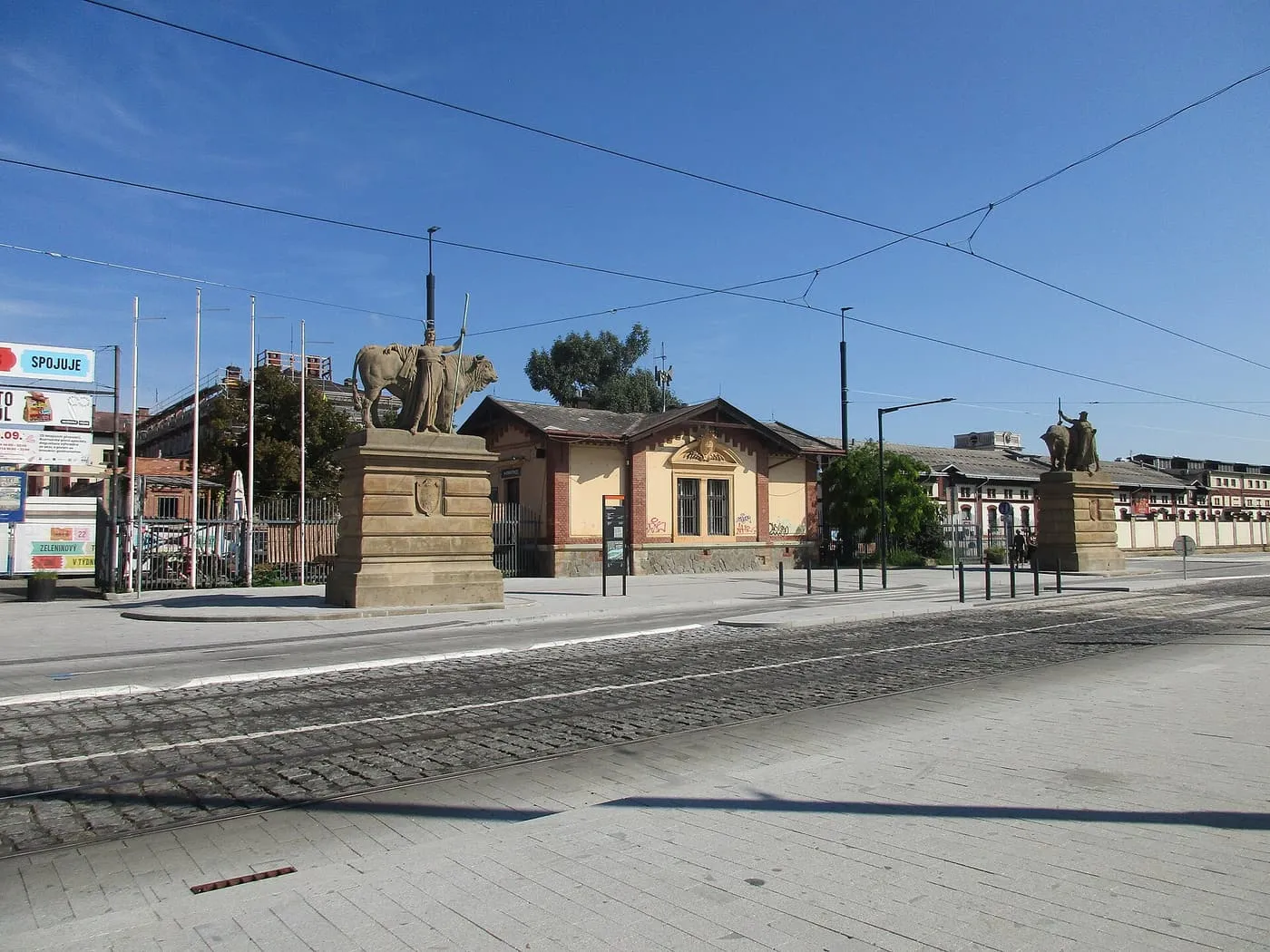 The sandstone gate piers of the Prague Market in Holešovice, topped with statues of figures with bulls, beside a small brick gatehouse and tram tracks