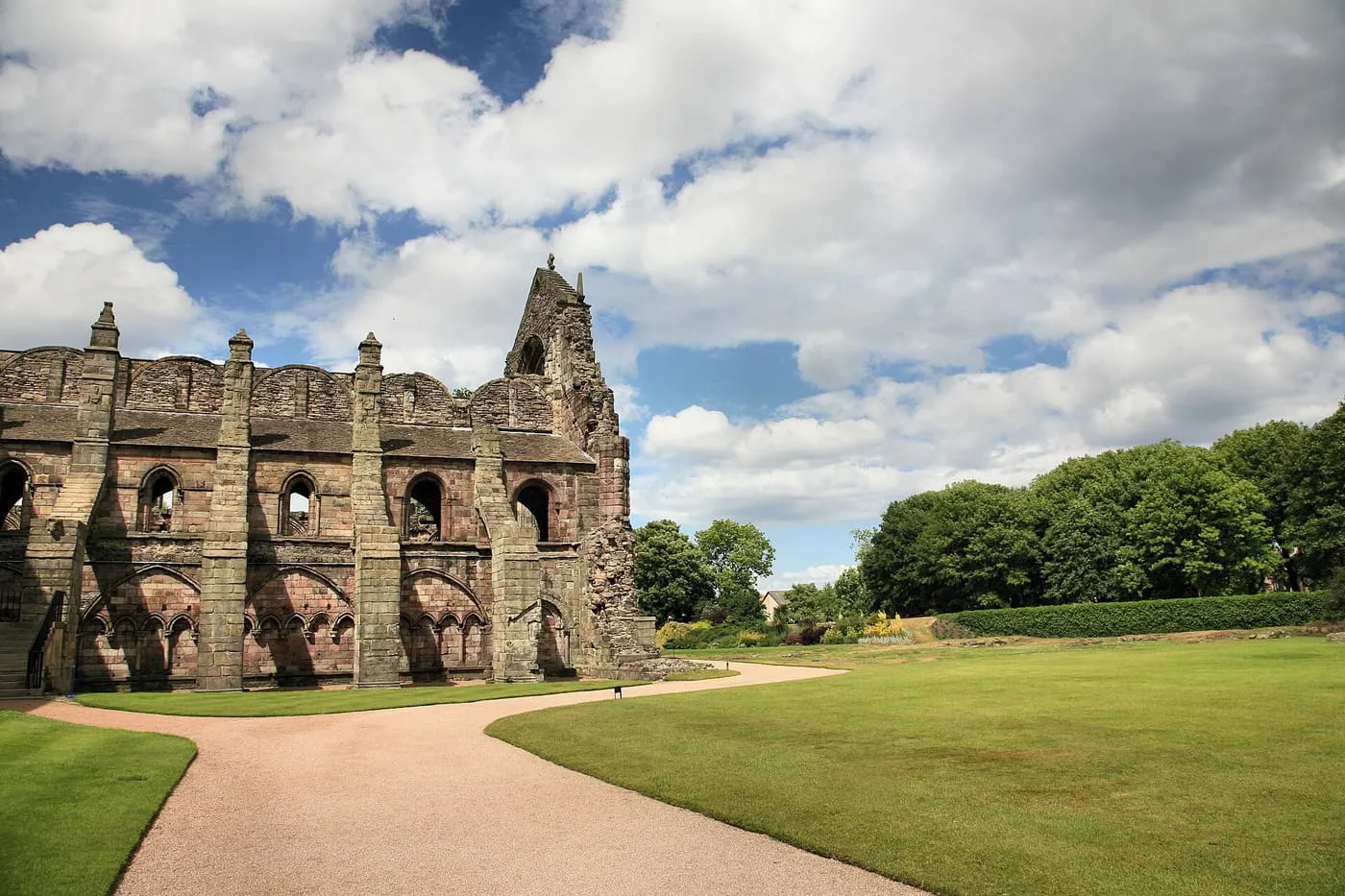 The roofless walls and arched windows of Holyrood Abbey beside a gravel path and a lawn