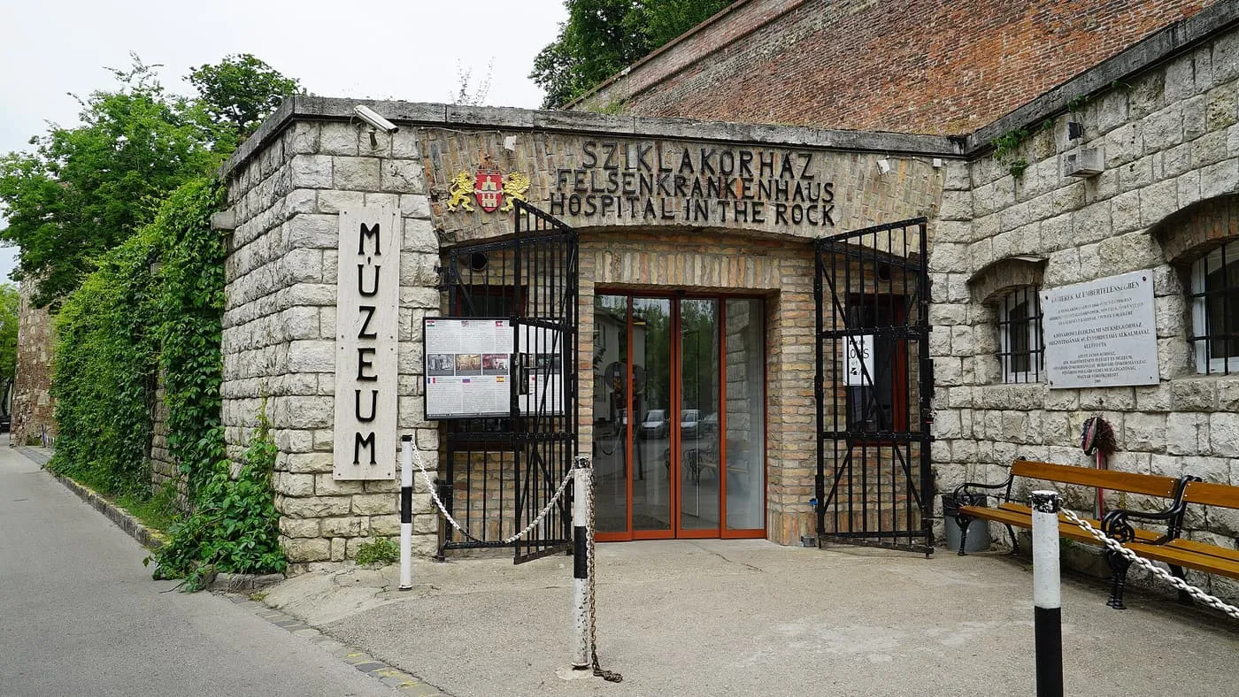 The stone entrance of the Hospital in the Rock museum in the castle wall, with "Sziklakórház Felsenkrankenhaus Hospital in the Rock" over the door and a bench beside it