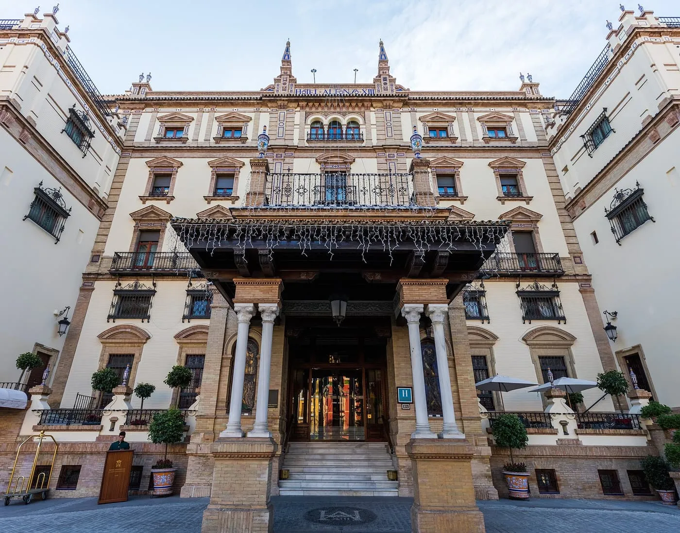 The entrance of the Hotel Alfonso XIII, a brick and stucco façade with a columned porch under a wooden canopy and potted trees at street level
