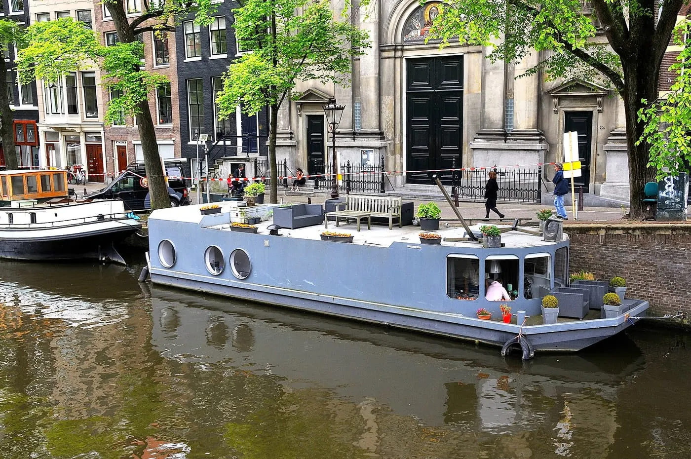 A grey houseboat with round portholes and a roof terrace moored at a brick quay on the Prinsengracht, in front of a church