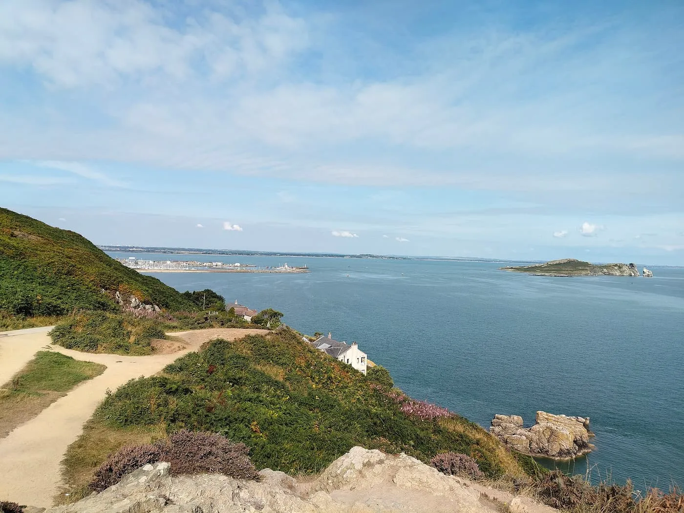 A sandy path along a heather and gorse cliff top above the sea, with Howth harbour and Ireland's Eye in the distance