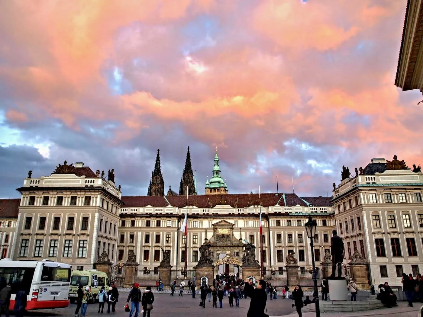 Hradčany Square at sunset, with the Castle's gate between two wings and the spires of St Vitus Cathedral above, under a pink sky