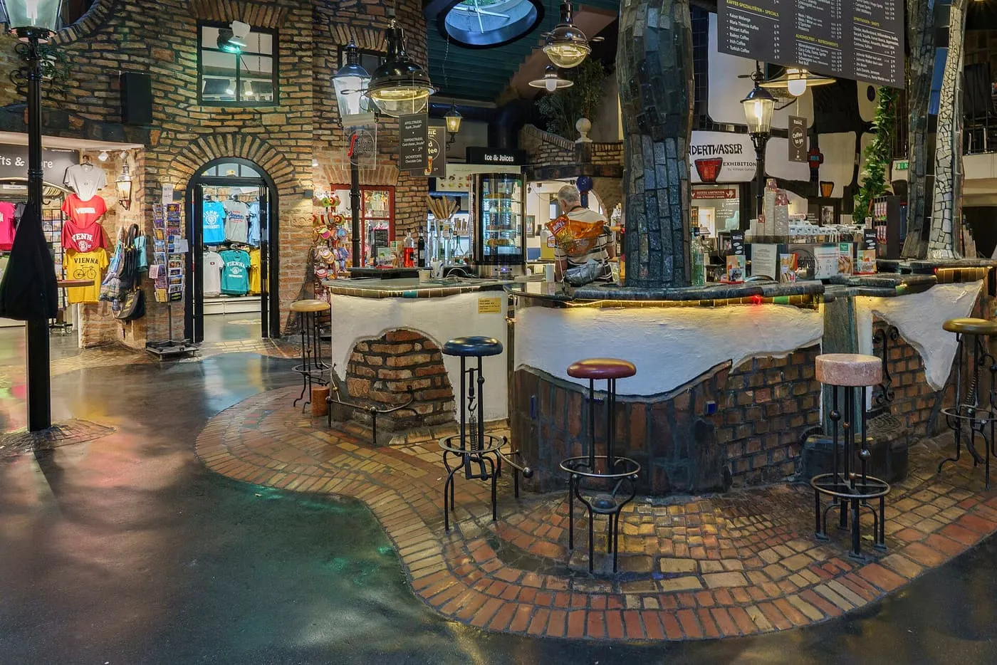 A café counter with an uneven tiled floor, a tree-trunk column and brick arches inside Hundertwasser Village, with a rack of T-shirts at left