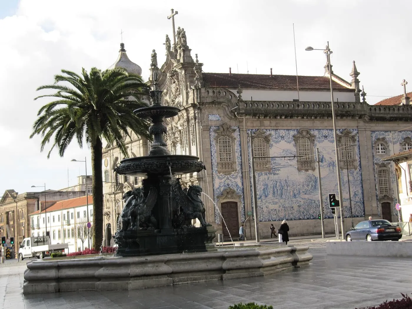 The long blue-and-white tiled side wall of the Igreja do Carmo beside a street, with a fountain and palm tree in the foreground