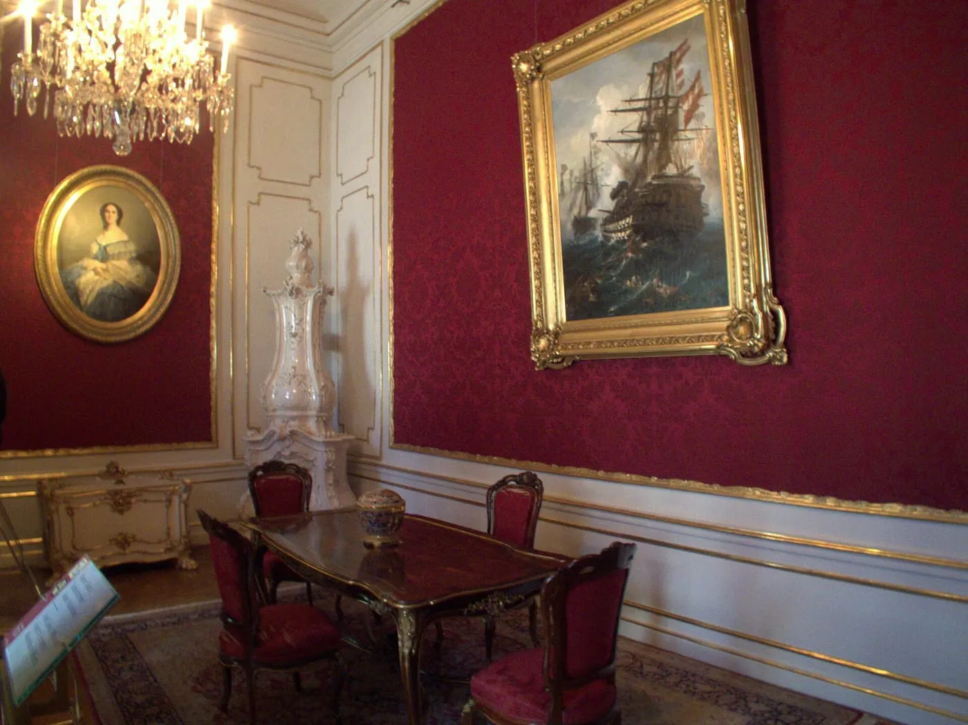 A room in the Imperial Apartments with crimson wall hangings, a gilt-framed ship painting, a white tiled stove and a desk with red chairs