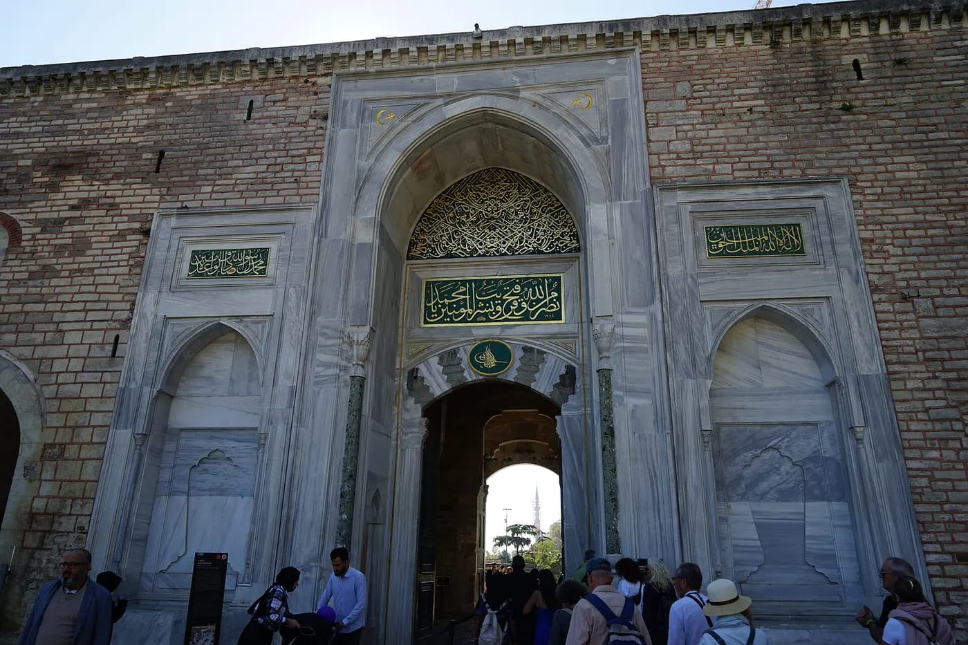 The marble Imperial Gate of Topkapı Palace in a brick wall, with gilded inscriptions over a pointed arch and visitors passing through
