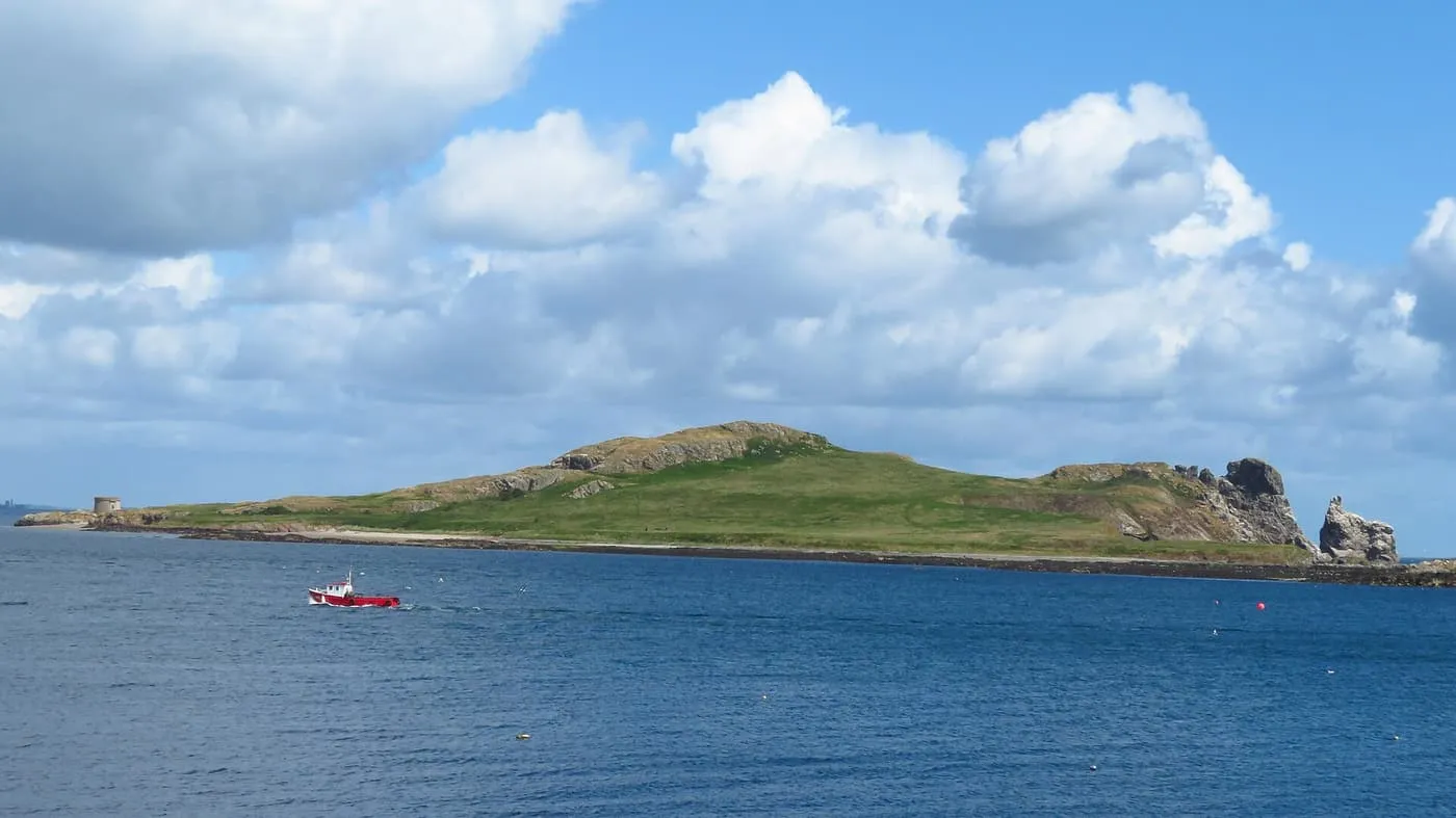 Ireland's Eye, a low green island with a small tower at its left end and sea stacks at the right, across blue water with a red fishing boat
