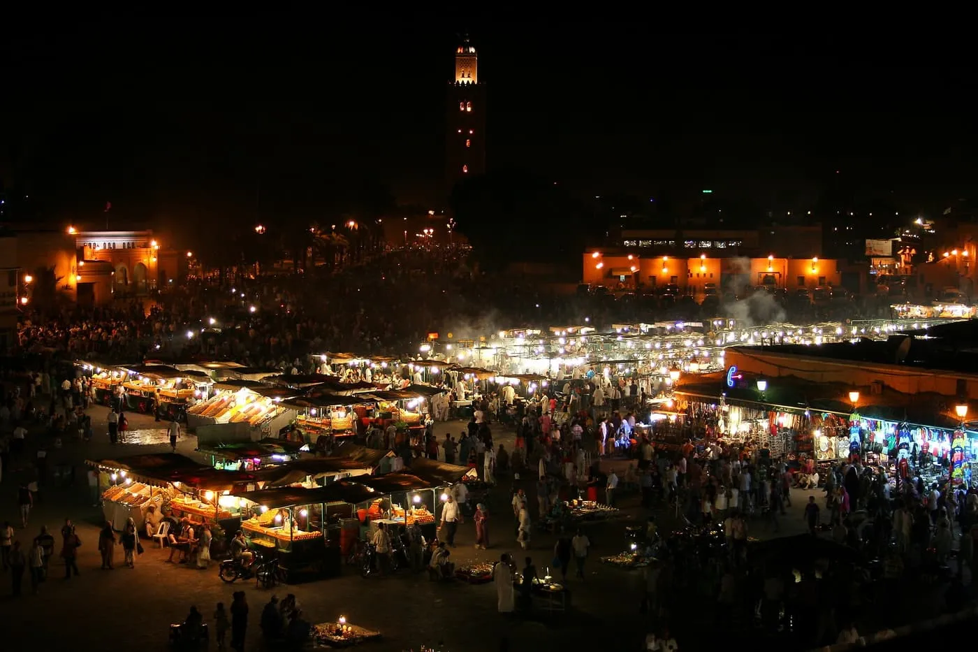 Jemaa el-Fnaa at night from above, with crowds among lit stalls, steam rising and the Koutoubia minaret lit in the dark behind