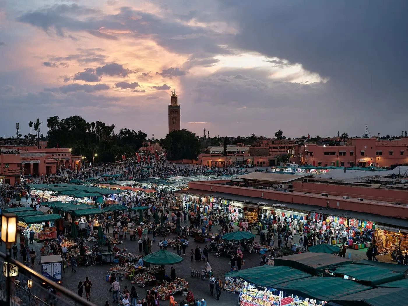 Jemaa el-Fnaa at dusk seen from above, with lit stalls and crowds in the square and the Koutoubia minaret against a pink and grey sky