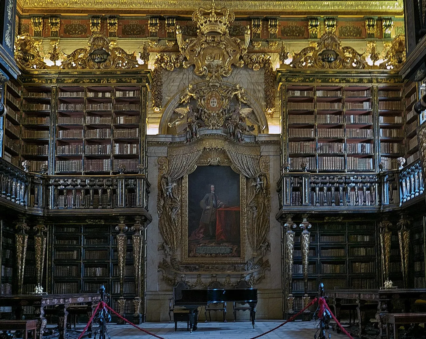 The Joanina Library, with gilded Baroque bookcases on two levels, a royal portrait and coat of arms at the far end, and red cords across the floor