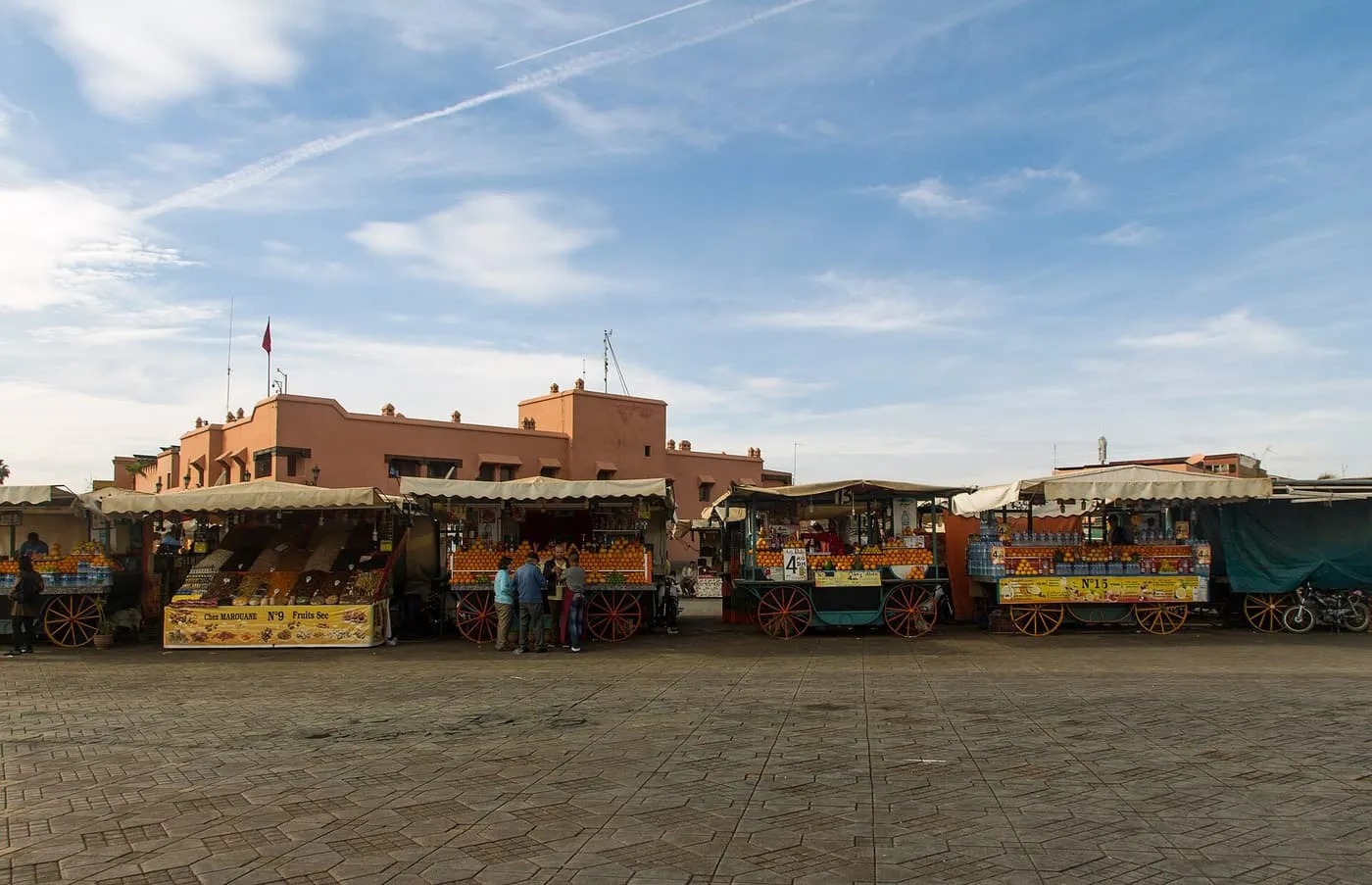 A row of orange-juice carts at the edge of a wide, empty Jemaa el-Fnaa under a blue sky