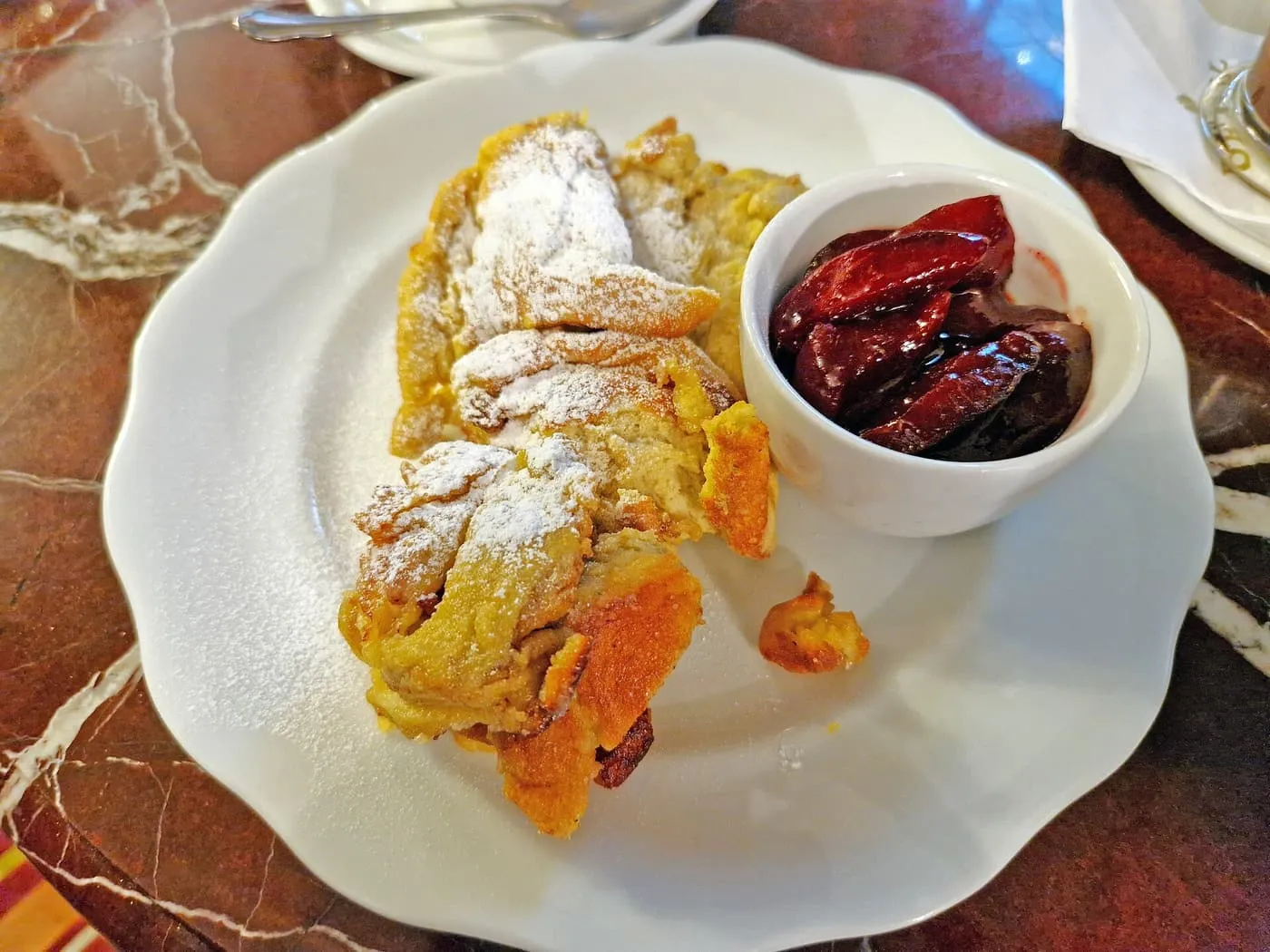Torn, sugar-dusted Kaiserschmarrn on a white plate with a small bowl of plum compote