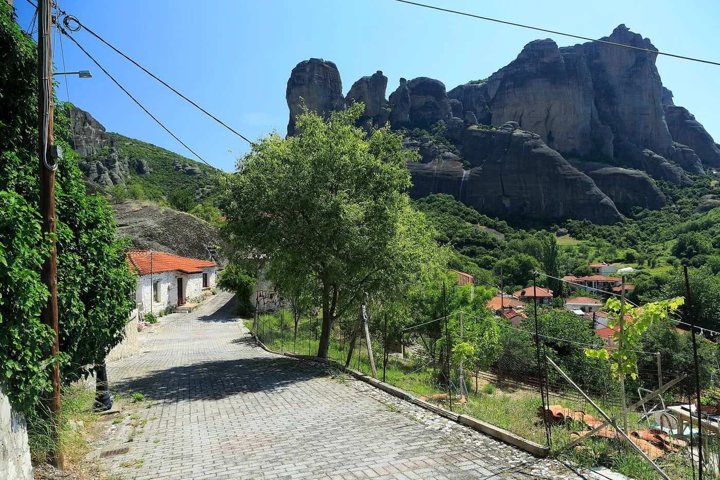 A cobbled street at the edge of Kalambaka with white houses and the Meteora rock pillars rising above the trees